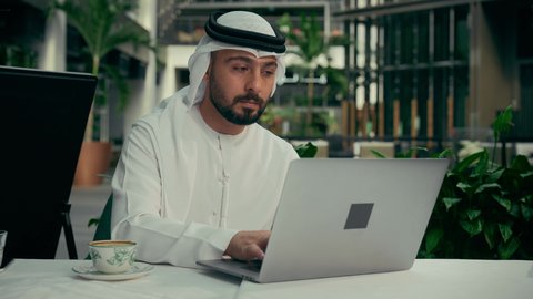 الاستمتاع بتناول فنجان من القهوة، جلسة خارجية في كافيه راقي في الإمارات العربية المتحدة, رجل عربي خليجي إماراتي يرتدي الكندورة والغترة يجلس في المقهى ويقوم بإنجاز العمل عن بعد عبر جهاز الحاسوب، استخدام الوسائل التقنية الحديثة والمتطورة