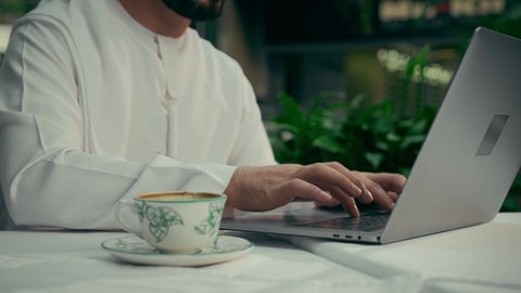 استخدام الوسائل التقنية الحديثة والمتطورة، الاستمتاع بتناول فنجان من القهوة، تصوير مقرب لرجل عربي خليجي إماراتي يرتدي الكندورة والغترة يجلس في المقهى ويقوم بإنجاز العمل عن بعد عبر جهاز الحاسوب، جلسة هادئة خارج المنزل، كافيه راقي في الإمارات العربية المتحدة