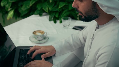 عرض شاشة لابتوب سوداء فارغة، الاستمتاع بتناول فنجان من القهوة، تصوير مقرب لرجل عربي خليجي إماراتي يرتدي الكندورة والغترة يجلس في المقهى ويقوم بإنجاز العمل عن بعد عبر جهاز الحاسوب، جلسة هادئة خارج المنزل، كافيه راقي في الإمارات العربية المتحدة