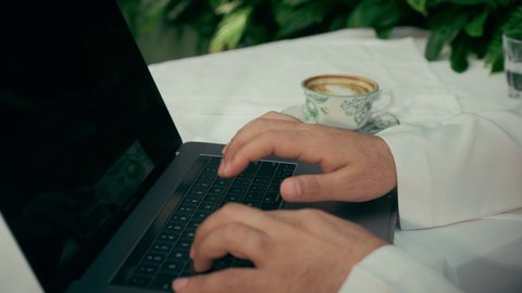 امتلاك مهارة الكتابة بسرعة، عرض شاشة لابتوب سوداء فارغة، الاستمتاع بتناول فنجان من القهوة، تصوير مقرب لأيدي رجل عربي خليجي إماراتي يجلس في المقهى ويقوم بالضغط على أزار لوحة المفاتيح، إنجاز العمل عن بعد عبر جهاز الحاسوب، كافيه راقي في الإمارات العربية المتحدة