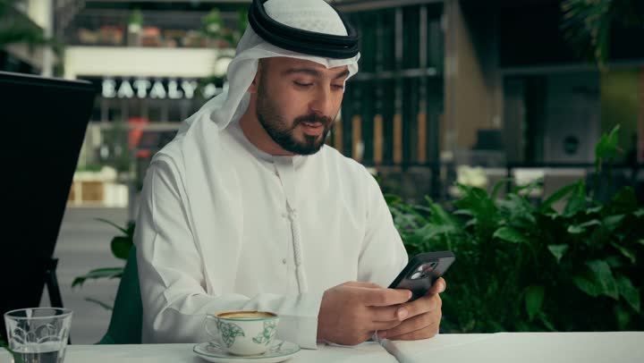 توثيق اللحظات السعيدة، الاستمتاع بالاجواء الهادئة خارج المنزل، رجل عربي خليجي اماراتي يرتدي الكندورة والغترة البيضاء يجلس في إحدى المقاهي الراقية يقوم بالتقاط صورة سيلفي تذكارية باستخدام الهاتف المحمول بإيماءات السرور، تناول فنجان من القهوة الساخنة