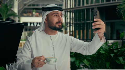 توثيق اللحظات السعيدة، الاستمتاع بالاجواء الهادئة خارج المنزل، رجل عربي خليجي اماراتي يرتدي الكندورة والغترة البيضاء يجلس في إحدى المقاهي الراقية يقوم بالتقاط صورة سيلفي تذكارية باستخدام الهاتف المحمول بإيماءات السرور، تناول فنجان من القهوة الساخنة