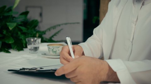 قراءة البنود الخاصة بالعمل، بناء شراكات واتفاقيات جديدة، احتساء القهوة الساخنة في الكافيه، الموافقة على العرض المالي، التحقق والتدقيق في شروط العقد، تصوير مقرب لأيدي رجل عربي خليجي اماراتي يجلس في إحدى المقاهي ويقوم بالتوقيع على العقود