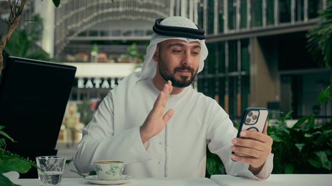 رفع اليد بإيماءات إلقاء التحية والسلام، مشاركة الأخبار والأحاديث الجيدة، الاستمتاع بالاجواء الهادئة خارج المنزل، رجل عربي خليجي اماراتي يرتدي الكندورة والغترة البيضاء يجلس في إحدى المقاهي الراقية ويقوم بإجراء مكالمة فيديو عبر الهاتف المحمول