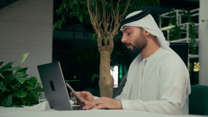 إيماءات الصدمة والخسارة، جلسة خارجية في كافيه راقي في الإمارات العربية المتحدة, تصوير من الجانب لرجل عربي خليجي إماراتي يرتدي الكندورة والغترة يجلس في المقهى ويقوم بإنجاز العمل عن بعد عبر جهاز الحاسوب، تصفح مواقع الانترنت باستخدام الهاتف المحمول