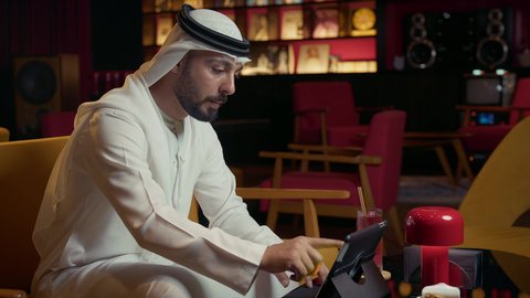 إيماءات الصدمة والخسارة بمشاهدة الأخبار السيئة، تناول كوب من العصير البارد، رجل عربي خليجي إماراتي يجلس في المقهى ويقوم بإنجاز العمل عن بعد عبر التابلت، كافيه راقي في الإمارات العربية المتحدة، تصفح مواقع التواصل الاجتماعي باستخدام الجهاز اللوحي