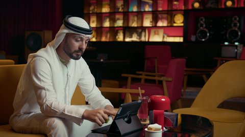 مفهوم الاستقبال والترحيب، إيماءات السعادة برؤية الأصدقاء، إلقاء التحيه والسلام, صداقة وتعارف وبناء علاقات اجتماعية، رجل عربي خليجي إماراتي يرتدي الكندورة والغترة يقابل صديقه في المقهى ويقوم بمصافحته، إنجاز العمل عن بعد عبر التابلت، كافيه راقي في الإمارات العربية