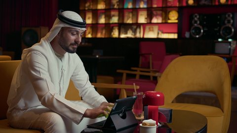 مفهوم الاستقبال والترحيب، إيماءات السعادة برؤية الأصدقاء، إلقاء التحيه والسلام, صداقة وتعارف وبناء علاقات اجتماعية، رجل عربي خليجي إماراتي يرتدي الكندورة والغترة يقابل صديقه في المقهى ويقوم بمصافحته، إنجاز العمل عن بعد عبر التابلت، كافيه راقي في الإمارات العربية