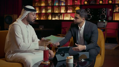 ايماءات الموافقة على المشاريع الجديدة، الحوار والنقاش بين الزملاء لإدارة الأعمال وتنظيمها، استخدام التقنيات الحديثة والمطورة، انجاز المهام عن بعد في الكافيه، رجل عربي خليجي اماراتي في اجتماع عمل مع عميل جديد يرتدي البدلة الرسمية، استخدام جهاز التابلت المحمول.