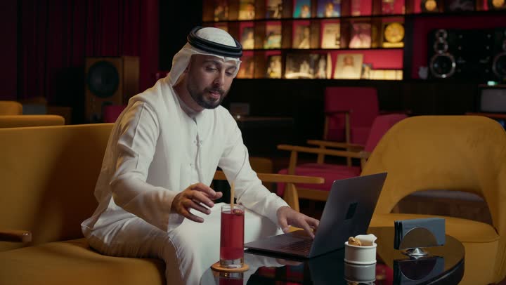 كافيه راقي في الإمارات العربية المتحدة، الاستمتاع بتناول المشروب البارد، استخدام الوسيلة التقنية الحديثة, رجل عربي خليجي إماراتي يرتدي الكندورة والغترة يجلس في المقهى ويقوم بإنجاز العمل عن بعد عبر جهاز الحاسوب