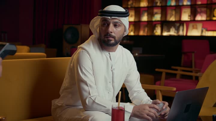 استخدام الوسيلة التقنية الحديثة في الدفع الالكتروني، سهولة الدفع الالي في الكافيهات الإماراتية, رجل عربي خليجي إماراتي يرتدي الكندورة والغترة يجلس في المقهى ويقوم بدفع الحساب عبر بطاقة البنك, الاستمتاع بتناول المشروب البارد ، إنجاز العمل عن بعد عبر جهاز الحاسوب