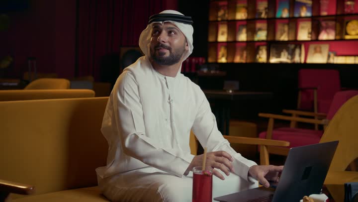 الاستمتاع بتناول المشروب البارد، استخدام الوسيلة التقنية الحديثة في الدفع الالكتروني، سهولة الدفع الالي في الكافيهات الإماراتية, رجل عربي خليجي إماراتي يرتدي الكندورة والغترة يجلس في المقهى ويقوم بدفع الحساب عبر بطاقة البنك, إنجاز العمل عن بعد عبر جهاز الحاسوب