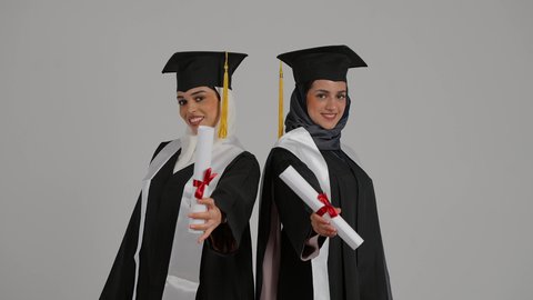 الاحتفال بالتخرج من الجامعة بإيماءات السعادة والفرح، التعليم الجامعي الاماراتي، أهمية الدراسة الجامعية والفرح بتحقيق النجاح،  تصوير مقرب لخريجتين عربيتين خليجيتين اماراتيتين مبتسمتين يرتديان ملابس التخرج وتمسكان وثيقة التفوق