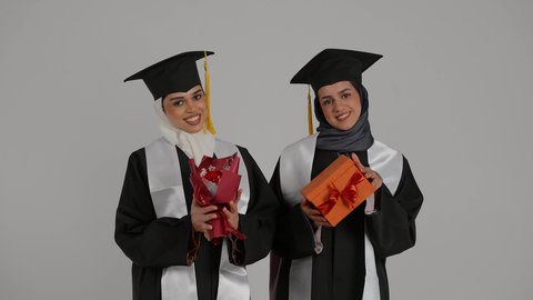 الشعور بالفخر والثقه بالنفس ، الاحتفال بالتخرج من الجامعة بإيماءات السعادة والفرح، التعليم الجامعي الاماراتي، أهمية الدراسة الجامعية والفرح بتحقيق النجاح،  تصوير مقرب لخريجتين عربيتين خليجيتين اماراتيتين مبتسمتين يرتديان ملابس التخرج وتمسكان هديه النجاح والورد