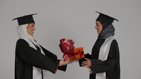 إيماءات السعادة والفرح، الشعور بالفخر والثقه بالنفس ، الاحتفال بالتخرج من الجامعة ، التعليم الجامعي الاماراتي، أهمية الدراسة الجامعية والفرح بتحقيق النجاح،  تصوير مقرب لخريجتين عربيتين خليجيتين اماراتيتين مبتسمتين يرتديان ملابس التخرج وتمسكان هديه النجاح والورد