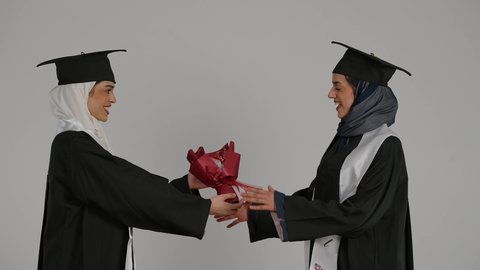 المناسبات والاحتفالات الجامعية, أهمية الدراسة الجامعية والفرح بتحقيق النجاح، التعليم الجامعي الاماراتي، إيماءات السعادة والفرح، الشعور بالفخر والثقه بالنفس ، الاحتفال بالتخرج من الجامعة ، تصوير مقرب لخريجتين عربيتين خليجيتين اماراتيتين مبتسمتين يرتديان ملابس التخرج وتمسكان ورد النجاح