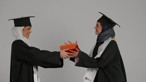 أهمية الدراسة الجامعية والفرح بتحقيق النجاح، التعليم الجامعي الاماراتي، إيماءات السعادة والفرح، الشعور بالفخر والثقه بالنفس ، الاحتفال بالتخرج من الجامعة ، تصوير مقرب لخريجتين عربيتين خليجيتين اماراتيتين مبتسمتين يرتديان ملابس التخرج وتمسكان هديه النجاح