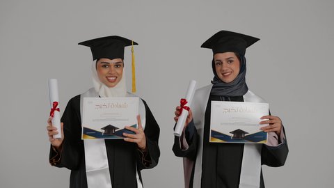 التعليم الجامعي الاماراتي، إيماءات السعادة والفرح، الشعور بالفخر والثقه بالنفس ، الاحتفال بالتخرج من الجامعة ، أهمية الدراسة الجامعية والفرح بتحقيق النجاح،  تصوير مقرب لخريجتين عربيتين خليجيتين اماراتيتين مبتسمتين يرتديان ملابس التخرج وتمسكان وثيقه النجاح