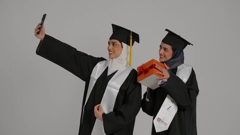 توثيق لحظات النجاح باستخدام الجوال ، المناسبات والاحتفالات الجامعية, إيماءات السعادة والفرح، أهمية الدراسة الجامعية والفرح بتحقيق النجاح، تصوير مقرب لخريجتين عربيتين خليجيتين اماراتيتين مبتسمتين يرتديان ملابس التخرج وتمسكان وثيقه النجاح ، احداهما تمسك هديه