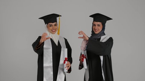 المناسبات والاحتفالات الجامعية, إيماءات السعادة والفرح، أهمية الدراسة الجامعية والفرح بتحقيق النجاح،  الاحتفال بالتخرج من الجامعة ، تصوير مقرب لخريجتين عربيتين خليجيتين اماراتيتين مبتسمتين يرتديان ملابس التخرج وتمسكان وثيقه النجاح ، ترفعان ايديهما على شكل قلب
