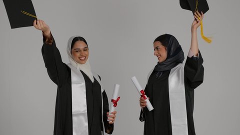 إيماءات السعادة والفرح، أهمية الدراسة الجامعية والفرح بتحقيق النجاح، التعليم الجامعي الاماراتي، المناسبات والاحتفالات الجامعية, الاحتفال بالتخرج من الجامعة ، تصوير مقرب لخريجتين عربيتين خليجيتين اماراتيتين مبتسمتين يرتديان ملابس التخرج وتمسكان وثيقه النجاح ، ترفعان قبعه التخرج