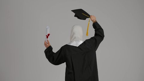 الشعور بالفخر والثقه بالنفس ، تحقيق حلم النجاح, إنهاء الدراسة الجامعية، مفهوم الاجتهاد والتفوق ، إيماءات الفرح والسرور، الاحتفال بالتخرج من الجامعة، تصوير مقرب من الخلف لطالبة عربية خليجية اماراتيه محجبة ترتدي روب التخرج ، ترفع وثيقه النجاح وقبعه التخرج بايديها