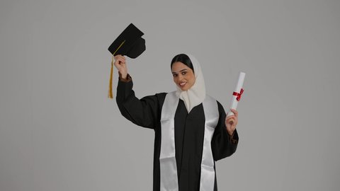 تحقيق حلم النجاح, إنهاء الدراسة الجامعية، مفهوم الاجتهاد والتفوق ، إيماءات الفرح والسرور، الشعور بالفخر والثقه بالنفس ، الاحتفال بالتخرج من الجامعة، تصوير مقرب لطالبة عربية خليجية اماراتيه محجبة ترتدي روب التخرج ، ترفع وثيقه النجاح وقبعه التخرج بايديها