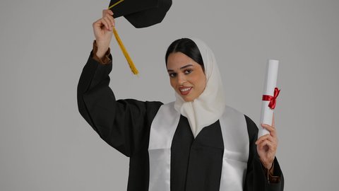 إنهاء الدراسة الجامعية، مفهوم الاجتهاد والتفوق ، إيماءات الفرح والسرور، الشعور بالفخر والثقه بالنفس ، تحقيق حلم النجاح, الاحتفال بالتخرج من الجامعة، تصوير مقرب لطالبة عربية خليجية اماراتيه محجبة ترتدي روب التخرج ، ترفع وثيقه النجاح وقبعه التخرج بايديها