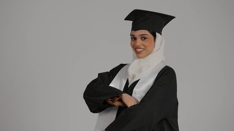مفهوم الاجتهاد والتفوق ، إيماءات الفرح والسرور، إنهاء الدراسة الجامعية، الشعور بالفخر والثقه بالنفس ، تحقيق حلم النجاح, الاحتفال بالتخرج من الجامعة، تصوير مقرب لطالبة عربية خليجية اماراتيه محجبة ترتدي روب التخرج والقبعة تقف بايدي متقاطعه