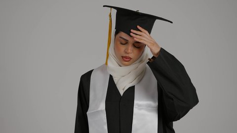 الحاجه للعلاج وطلب المساعده ، إنهاء الدراسة الجامعية، تحقيق حلم النجاح, الاحتفال بالتخرج من الجامعة، مفهوم الاجتهاد والتفوق ، تصوير مقرب لطالبة عربية خليجية اماراتيه محجبة ترتدي روب التخرج والقبعة تضع يدها على رأسها بإيماءات الدوار و الألم