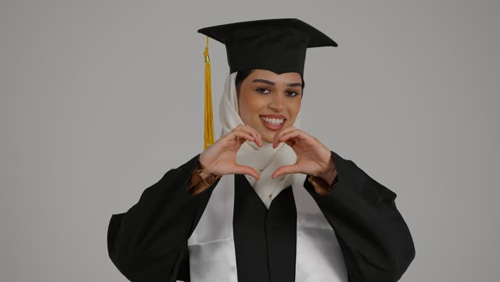 إنهاء الدراسة الجامعية، تحقيق حلم النجاح, الاحتفال بالتخرج من الجامعة، مفهوم الاجتهاد والتفوق، رفع وثيقه النجاح بتعابير الفخر، الشعور بالثقة بالنفس ، تصوير مقرب لطالبة عربية خليجية اماراتيه محجبة ترتدي روب التخرج ، ترفع يديها للاعلى على شكل قلب