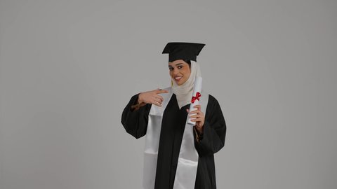 مفهوم الاجتهاد والتفوق، الاشاره الى وثيقه النجاح بتعابير الفخر ، تحقيق حلم النجاح, الشعور بالثقة بالنفس ، الاحتفال بإنهاء الدراسة الجامعية، تصوير مقرب لطالبة عربية خليجية اماراتيه محجبة ترتدي روب التخرج ، ترفع وثيقه النجاح بايماءات السرور