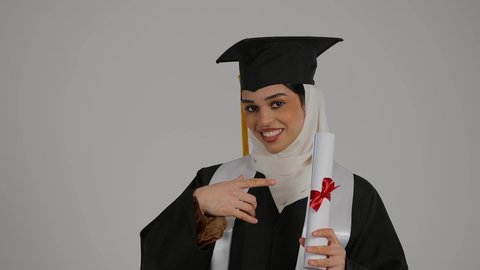 الاشاره الى وثيقه النجاح بتعابير الفخر ، تحقيق حلم النجاح, الشعور بالثقة بالنفس ، الاحتفال بإنهاء الدراسة الجامعية، مفهوم الاجتهاد والتفوق، تصوير مقرب لطالبة عربية خليجية اماراتيه محجبة ترتدي روب التخرج ، ترفع وثيقه النجاح بايماءات السرور