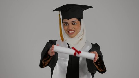 ايماءات الفخر والسرور بتحقيق حلم النجاح, الشعور بالثقة بالنفس والفخر، الاحتفال بإنهاء الدراسة الجامعية، مفهوم الاجتهاد والتفوق، تصوير مقرب لطالبة عربية خليجية اماراتيه محجبة ترتدي روب التخرج ، ترفع وثيقه النجاح بايماءات السرور