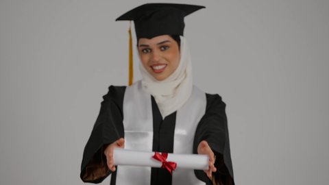 الشعور بالثقة بالنفس والفخر، الاحتفال بإنهاء الدراسة الجامعية، مفهوم الاجتهاد والتفوق، الفرح بتحقيق النجاح، تصوير مقرب لطالبة عربية خليجية اماراتيه محجبة ترتدي روب التخرج ، ترفع وثيقه النجاح بايماءات السرور