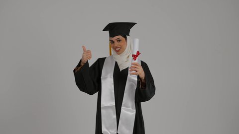 مفهوم الاجتهاد والتفوق، تعابير الجودة والامتياز والموافقه, الشعور بالثقة بالنفس والفخر، الفرح بتحقيق النجاح، تصوير مقرب لطالبة عربية خليجية اماراتيه محجبة ترتدي روب التخرج ، ترفع وثيقه النجاح بايماءات السرور
