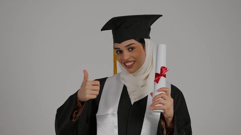 تعابير الجودة والامتياز والموافقه, الشعور بالثقة بالنفس والفخر، الفرح بتحقيق النجاح، مفهوم الاجتهاد والتفوق، تصوير مقرب لطالبة عربية خليجية اماراتيه محجبة ترتدي روب التخرج ، ترفع وثيقه النجاح بايماءات السرور