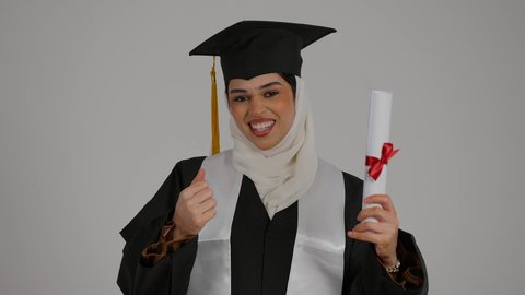 رفع قبضه اليد بايماءات السعاده , الشعور بالثقة بالنفس والفخر، الفرح بتحقيق النجاح، مفهوم الاجتهاد والتفوق، تصوير مقرب لطالبة عربية خليجية اماراتيه محجبة ترتدي روب التخرج ، ترفع وثيقه النجاح بايماءات السرور