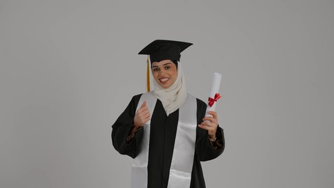 احتفالات التخرج الجامعية, الشعور بالثقة بالنفس والفخر، الفرح بتحقيق النجاح، تعابير السرور والفرح ، مفهوم الاجتهاد والتفوق، تصوير مقرب من الجانب لطالبة عربية خليجية اماراتيه محجبة ترتدي روب التخرج ، ترفع وثيقه النجاح بايماءات السعاده