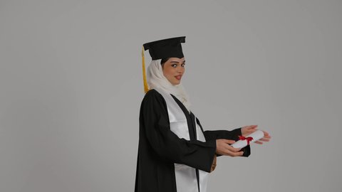 الشعور بالثقة بالنفس والفخر، أهمية الدراسة الجامعية والفرح بتحقيق النجاح،الاحتفالات والمناسبات الجامعية, تعابير السرور ، مفهوم الاجتهاد والتفوق، تصوير مقرب من الجانب لطالبة عربية خليجية اماراتيه محجبة ترتدي روب التخرج ، ترفع وثيقه النجاح بايماءات السعاده