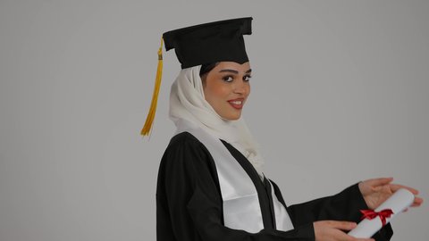 أهمية الدراسة الجامعية والفرح بتحقيق النجاح، ايماءات الجودة والامتياز ،الاحتفالات والمناسبات الجامعية, تعابير السرور ، الشعور بالثقة بالنفس والفخر، مفهوم الاجتهاد والتفوق، تصوير مقرب من الجانب لطالبة عربية خليجية اماراتيه محجبة ترتدي روب التخرج ، ترفع وثيقه النجاح بايماءات السعاده