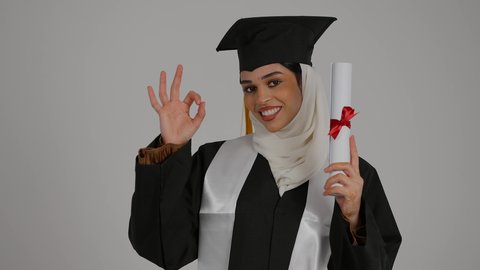 لحظة تاريخية في حياة الخريجين، ايماءات الجودة والامتياز ،الاحتفالات والمناسبات الجامعية, تعابير السرور و الفرح لتحقيق النجاح، الشعور بالثقة بالنفس والفخر، مفهوم الاجتهاد والتفوق، تصوير مقرب لطالبة عربية خليجية اماراتيه محجبة ترتدي روب التخرج ، ترفع وثيقه النجاح بايماءات السعاده