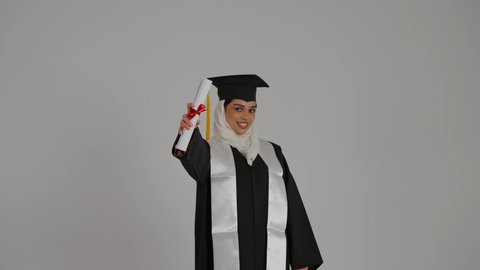 الشعور بالثقة بالنفس والفخر، جودة التعليم في المملكة ، الاحتفالات والمناسبات الجامعية,  تحقيق حلم النجاح ، مفهوم الاجتهاد والتفوق،تصوير مقرب لطالبة عربية خليجية اماراتيه محجبة ترتدي روب التخرج ، ترفع وثيقه النجاح بايماءات السعاده