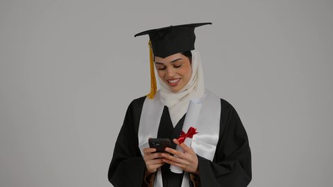 دمج التقنيات الحديثه فى الحياه اليوميه ، الاحتفالات والمناسبات الجامعية, تحقيق حلم النجاح ، الشعور بالثقة بالنفس والفخر، جودة التعليم في المملكة ، مفهوم الاجتهاد والتفوق،تصوير مقرب لطالبة عربية خليجية اماراتيه محجبة ترتدي روب التخرج ، تحمل بيدها الجوال