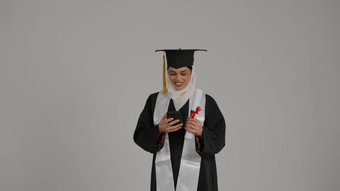 اجراء مكالمه هاتفيه ، الاحتفالات والمناسبات الجامعية, تحقيق حلم النجاح ، الشعور بالثقة بالنفس والفخر، جودة التعليم في المملكة ، مفهوم الاجتهاد والتفوق،تصوير مقرب لطالبة عربية خليجية اماراتيه محجبة ترتدي روب التخرج ، تحمل بيدها وثيقه النجاح وقبعة التخرج