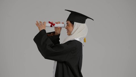 الاحتفالات والمناسبات الجامعية, النظر من خلال شهاده التخرج ، تحقيق حلم النجاح ، الشعور بالثقة بالنفس والفخر، جودة التعليم في المملكة ، مفهوم الاجتهاد والتفوق،تصوير مقرب لطالبة عربية خليجية اماراتيه محجبة ترتدي روب التخرج ، تحمل بيدها وثيقه النجاح وقبعة التخرج