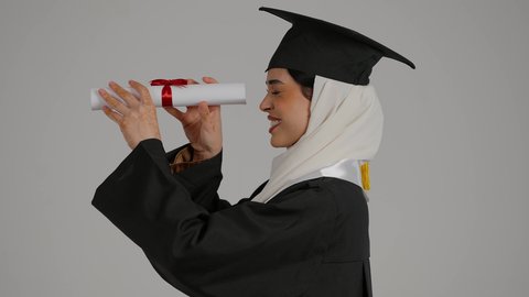 النظر من خلال شهاده التخرج ، تحقيق حلم النجاح ، الشعور بالثقة بالنفس والفخر، جودة التعليم في المملكة ، الاحتفالات والمناسبات الجامعية, مفهوم الاجتهاد والتفوق،تصوير مقرب لطالبة عربية خليجية اماراتيه محجبة ترتدي روب التخرج ، تحمل بيدها وثيقه النجاح وقبعة التخرج