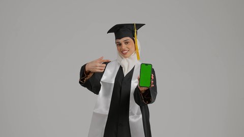 عرض شاشه خضراء كروما ، الاحتفالات والمناسبات الجامعية, مفهوم الاجتهاد والتفوق، الشعور بالثقة بالنفس والفخر، تصوير مقرب لطالبة عربية خليجية اماراتيه محجبة ترتدي روب وقبعة التخرج ،تحمل بيدها الهاتف المحمول ، تضع وشاح علم الامارات على كتفها، إنهاء الدراسة الجامعية، جودة التعليم في المملكة