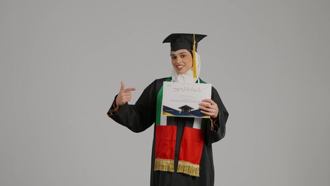 الاشاره الى شهادة التخرج بايماءات الفخر ، تصوير مقرب لطالبة عربية خليجية اماراتيه محجبة ترتدي عباءه وقبعة التخرج ، تضع وشاح علم الامارات على كتفها، تحمل بيدها وثيقه النجاح، الاحتفالات والمناسبات الجامعية, مفهوم الاجتهاد والتفوق، إنهاء الدراسة الجامعية، الشعور بالفخر والثقة بالنفس، جودة التعليم في المملكة