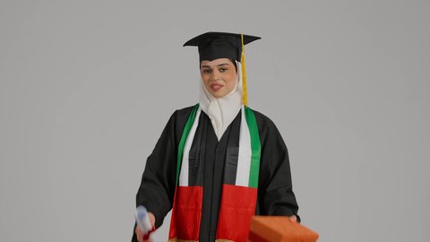 جودة التعليم في المملكة، النظر الى الكاميرا بايماءات السرور،الاحتفالات و المناسبات الجامعية,  الشعور بالفخر والثقة بالنفس، إنهاء الدراسة الجامعية، مفهوم الاجتهاد والتفوق، تصوير مقرب لطالبة عربية خليجية اماراتيه محجبة ترتدي عباءه وقبعة التخرج تحمل بيدها هدية النجاح والوثيقه